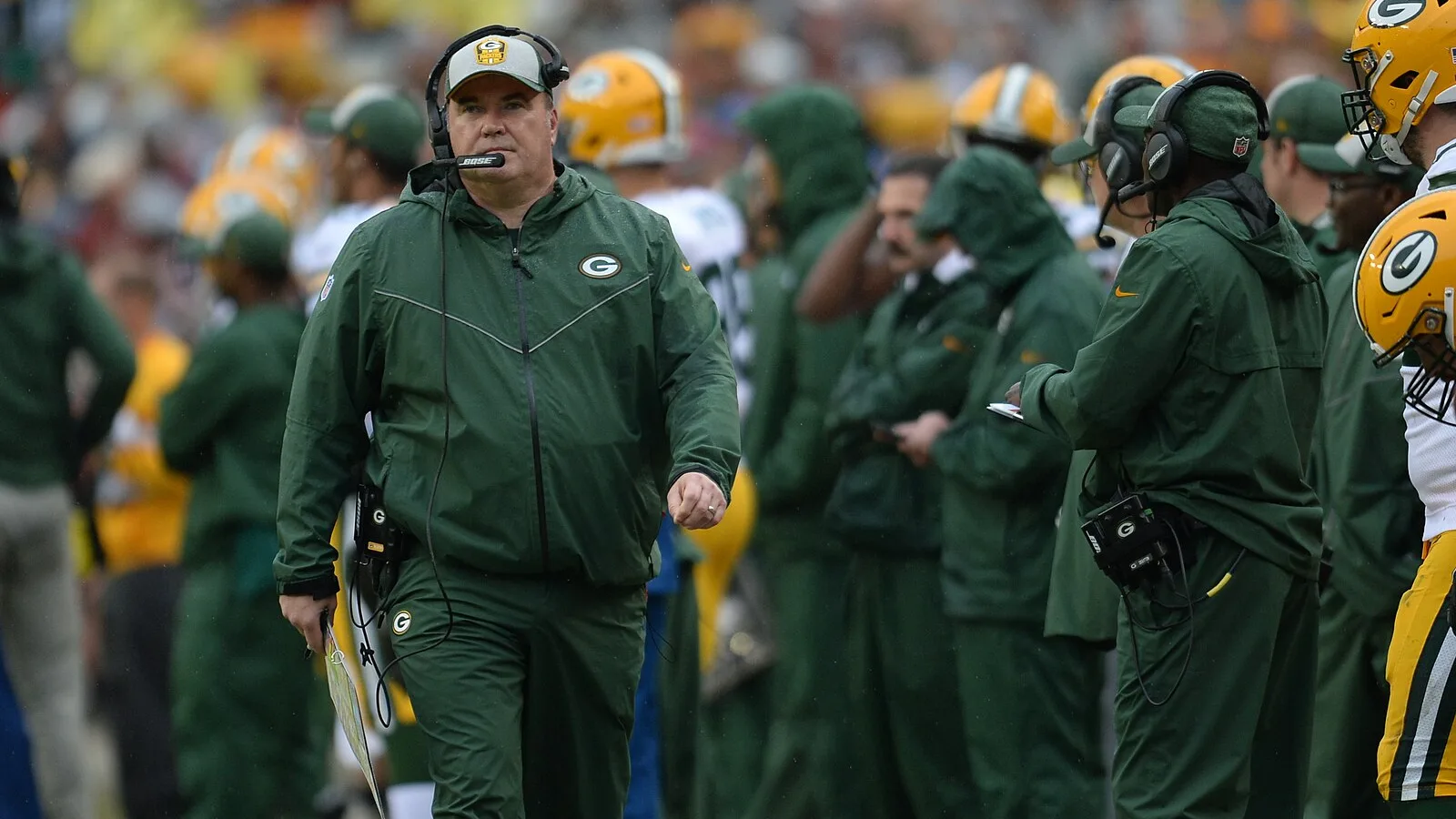Mike McCarthy in a green Packers jacket and headset walking the sideline